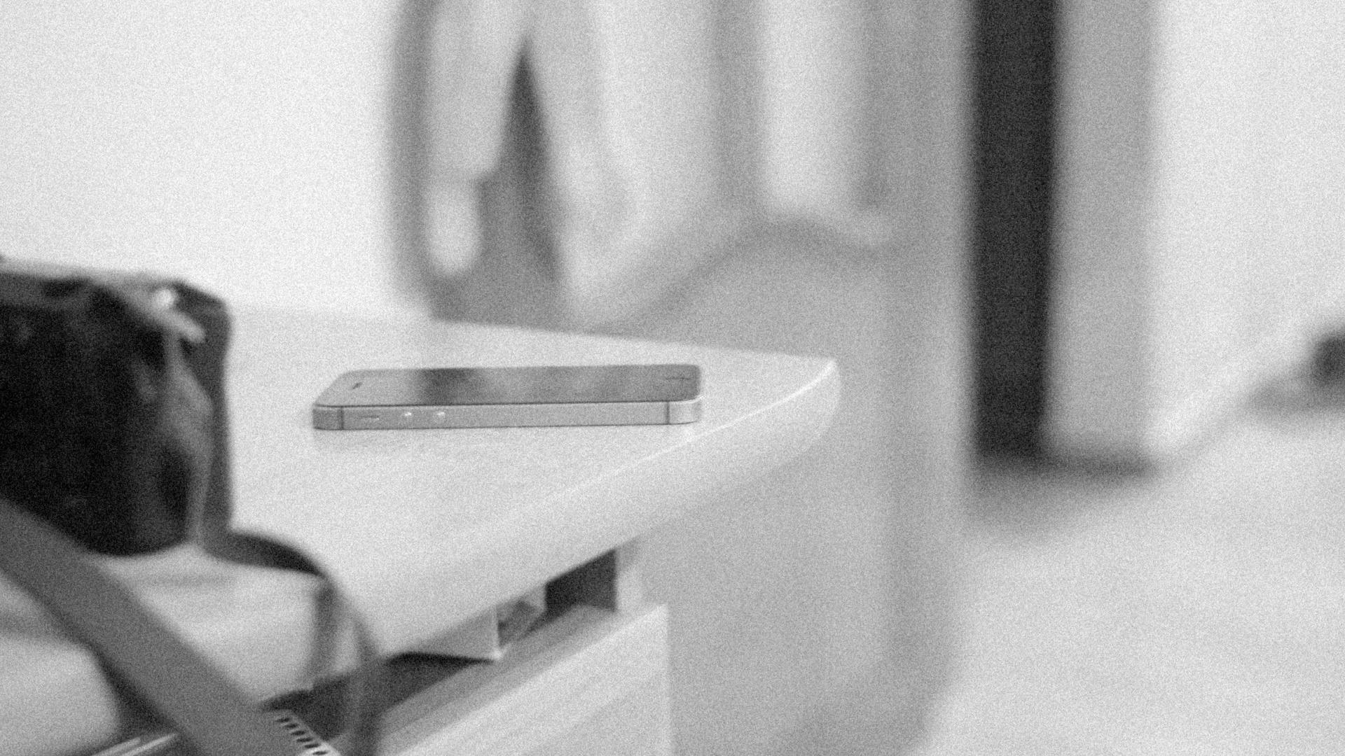 A smartphone lying flat on the corner of a white table, a dark bag out of focus beside it, in black and white