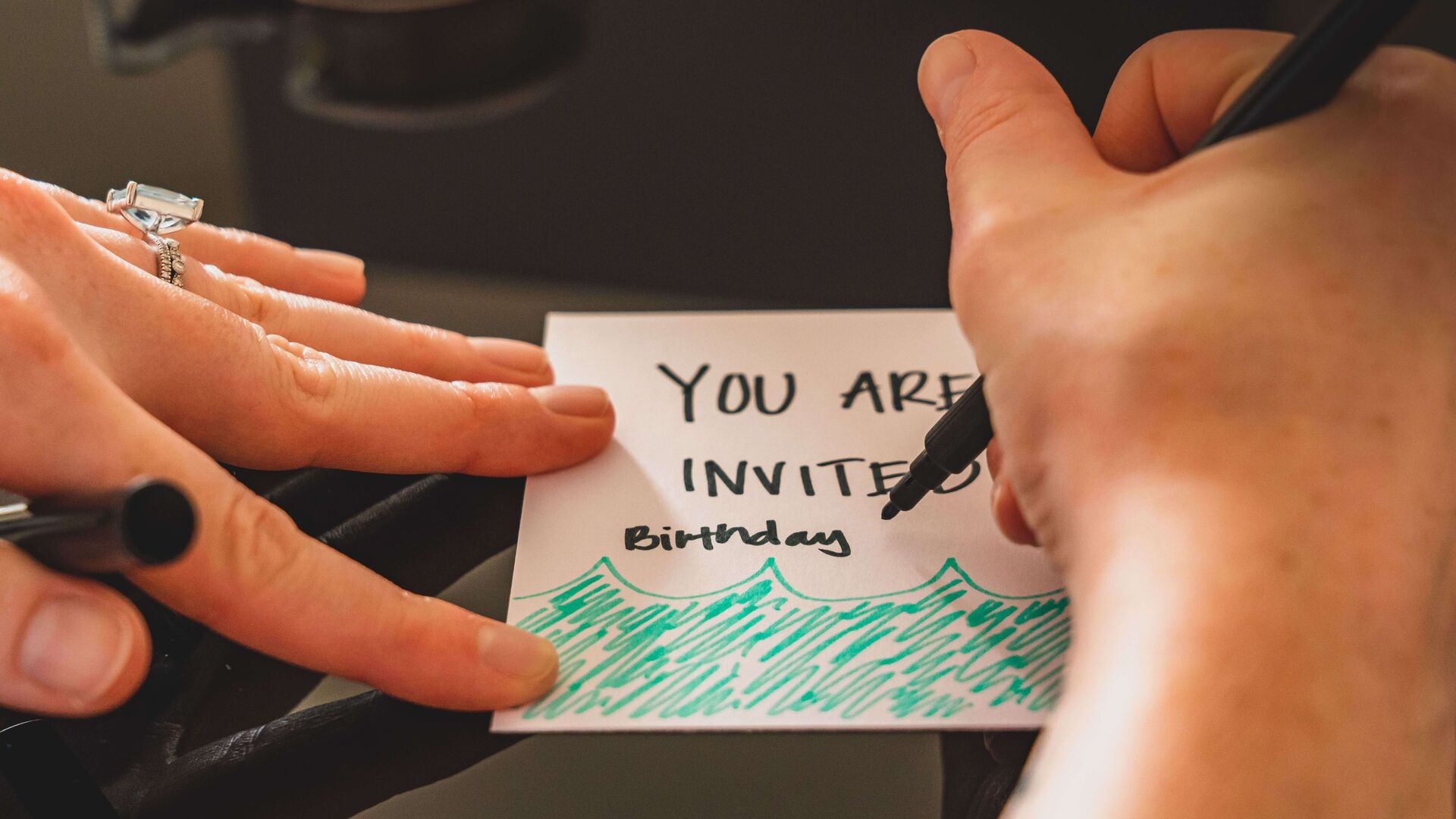 Hands writing a card that says "You are invited"